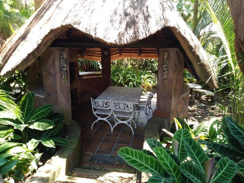 A rustic outdoor thatched lapa area featuring a built-in braai, tiled flooring, and masonry pillars, set within a lush garden environment.