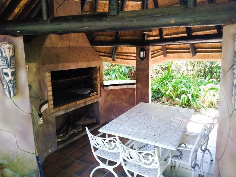 Covered outdoor patio area featuring a built-in braai, traditional thatched roof, and tiled flooring, overlooking a lush garden setting.