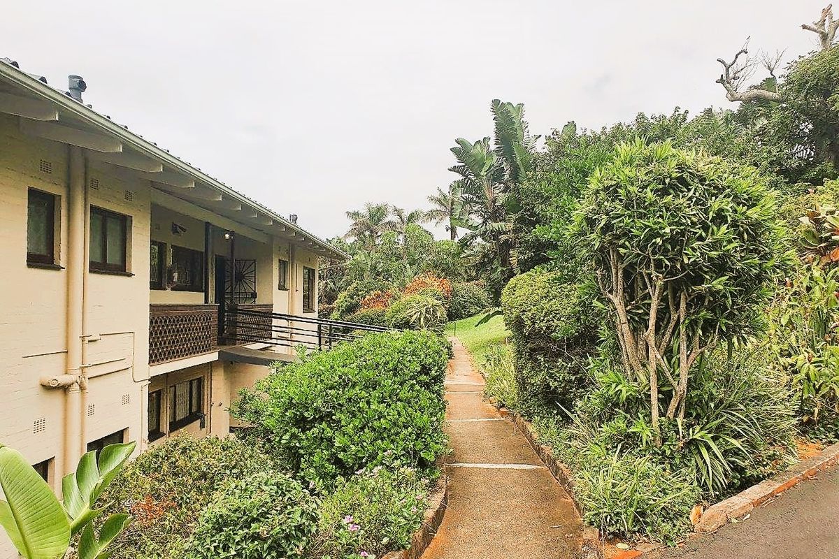 A view of the apartment building exterior featuring a concrete walkway leading through a lush, established garden area.