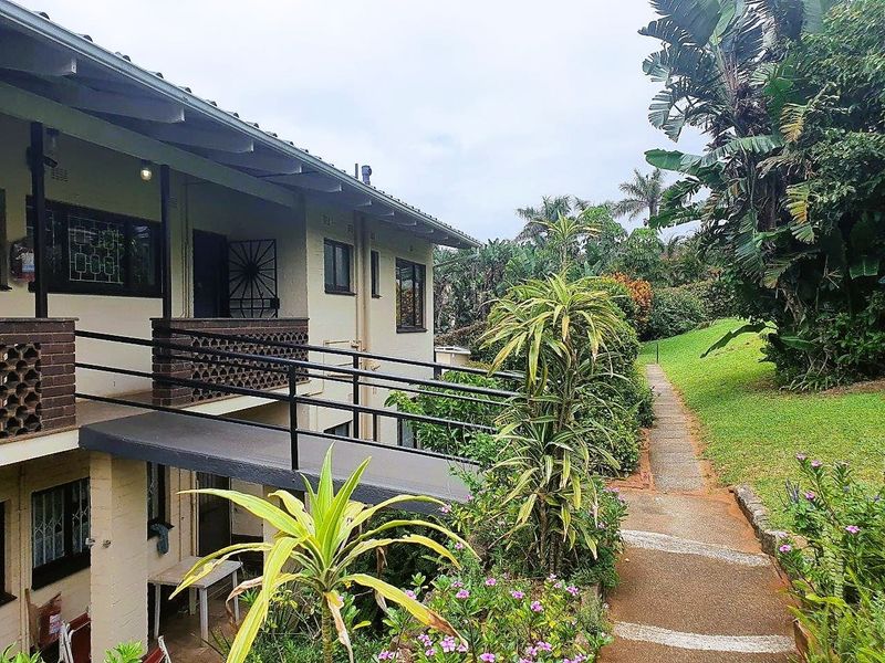 Rear exterior view of the apartment building featuring a raised walkway, metal balustrades, and access to the landscaped garden area.