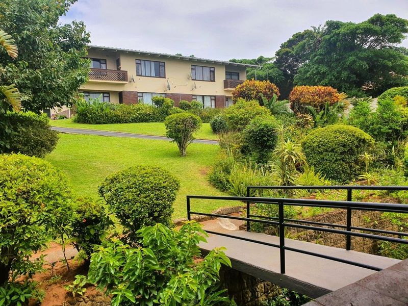 A view of the apartment building exterior featuring landscaped gardens, a pedestrian bridge, and private balconies.