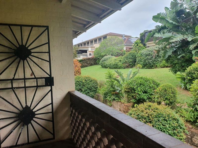 A covered balcony featuring a security gate and brick balustrade, overlooking a lush, well-maintained communal garden area.