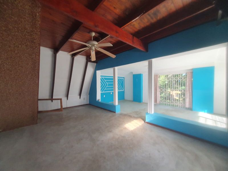 Spacious open-plan living area featuring exposed timber ceiling beams, a ceiling fan, and a security gate, offering a blank canvas for renovation.