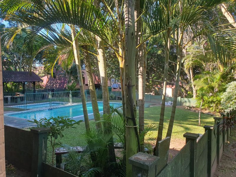 A communal outdoor area featuring a swimming pool, surrounding lawn, and a covered gazebo structure within a landscaped garden setting.