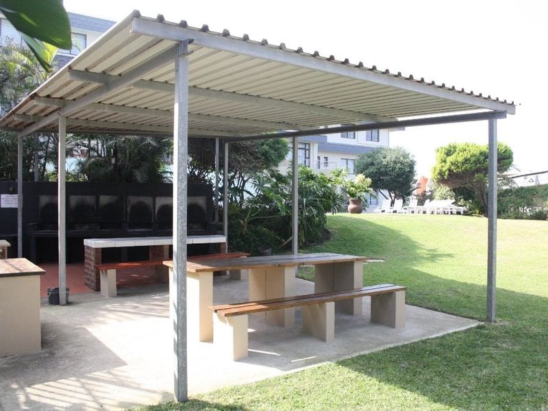 Covered outdoor entertainment area featuring a built-in brick braai, concrete seating, and a corrugated metal roof set within a communal garden space.