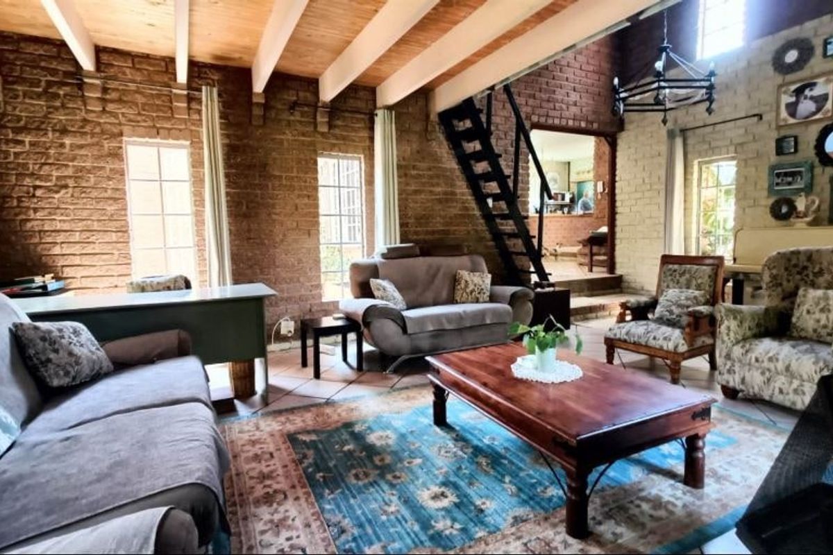 Spacious living room featuring rustic exposed brick walls, timber ceiling beams, and terracotta flooring, with a metal staircase leading to an upper level.