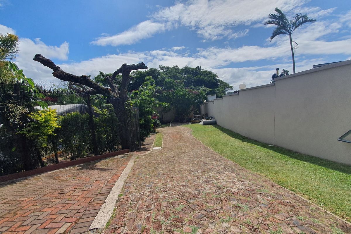 A view of the rear exterior area featuring a brick-paved driveway, a long perimeter boundary wall, and established garden landscaping.