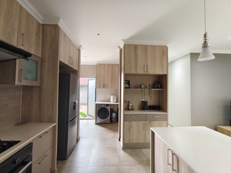 Modern kitchen featuring light wood-grain cabinetry, stone countertops, integrated appliances, and a seamless transition to a rear utility area with laundry connections.