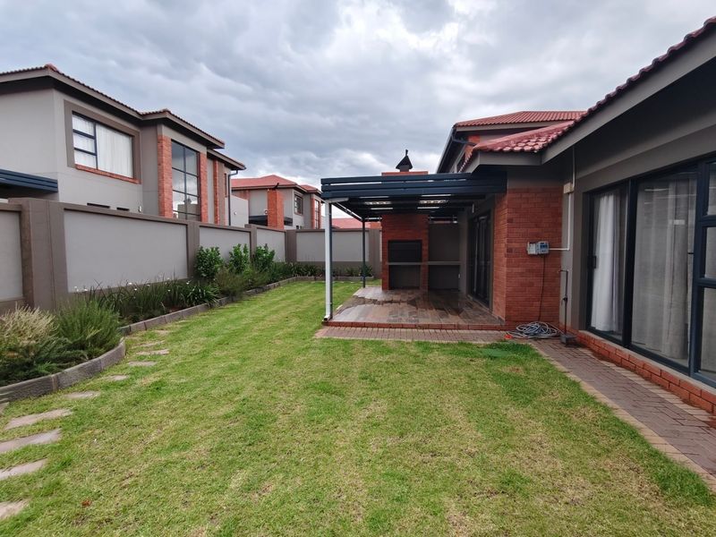 Rear garden featuring a covered patio with a built-in braai, brick perimeter walls, and a well-maintained lawn area.