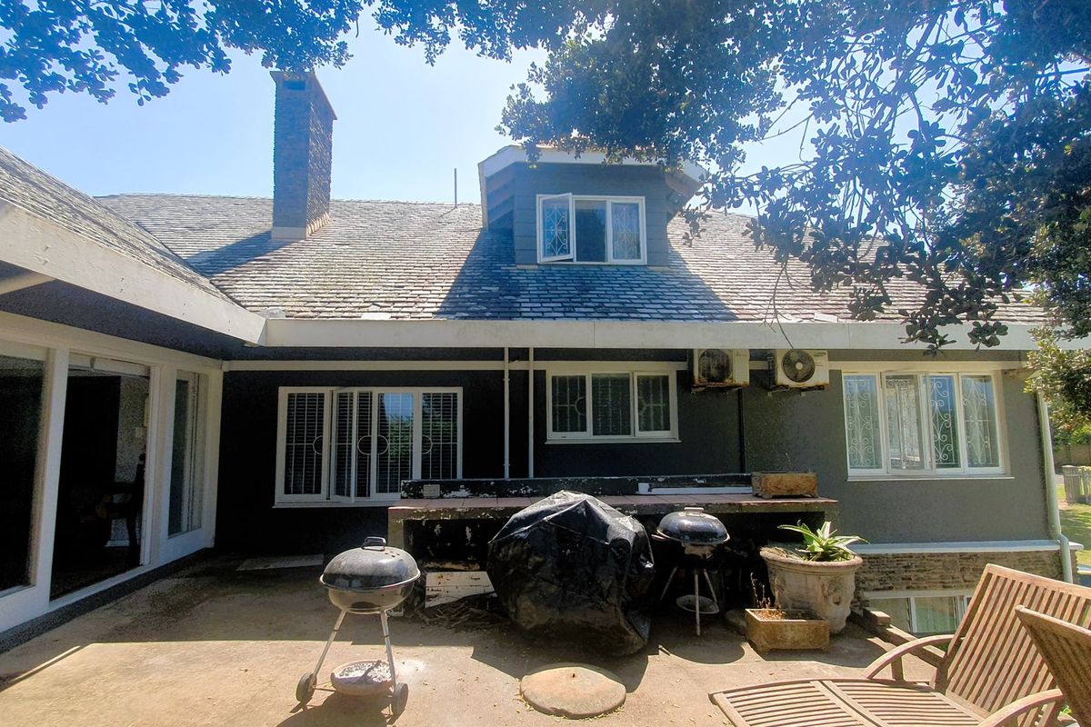 Rear exterior view of the house featuring a concrete patio, built-in braai area, slate roof, and established garden surroundings.