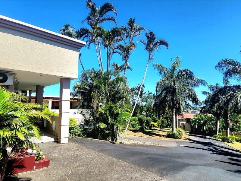 Front exterior view of the property featuring a paved driveway, covered entrance area, and established garden with mature palm trees.