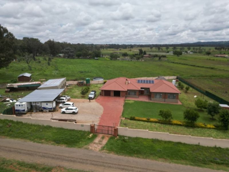 Aerial view of a residential property featuring a main house with solar panels, a paved driveway, perimeter walling, and multiple outbuildings on a large plot.