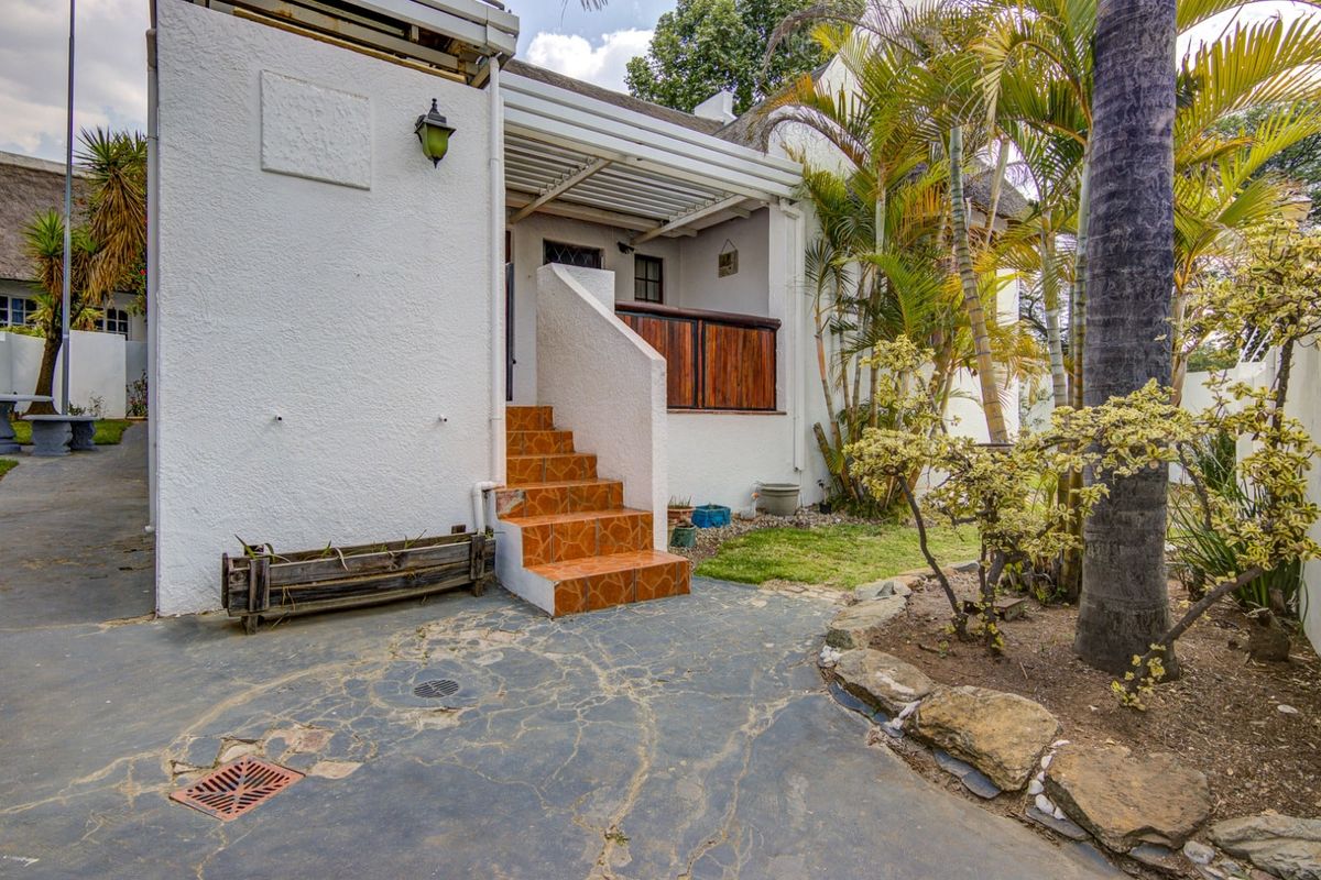 Rear exterior view featuring a covered patio, tiled steps, and a landscaped garden area with mature trees and paved pathways.