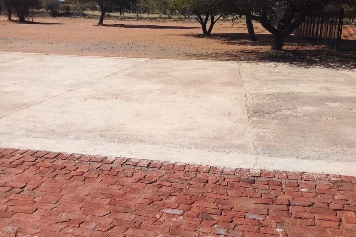 An outdoor area featuring a combination of brick and concrete paving, leading to an open farm landscape with mature trees.