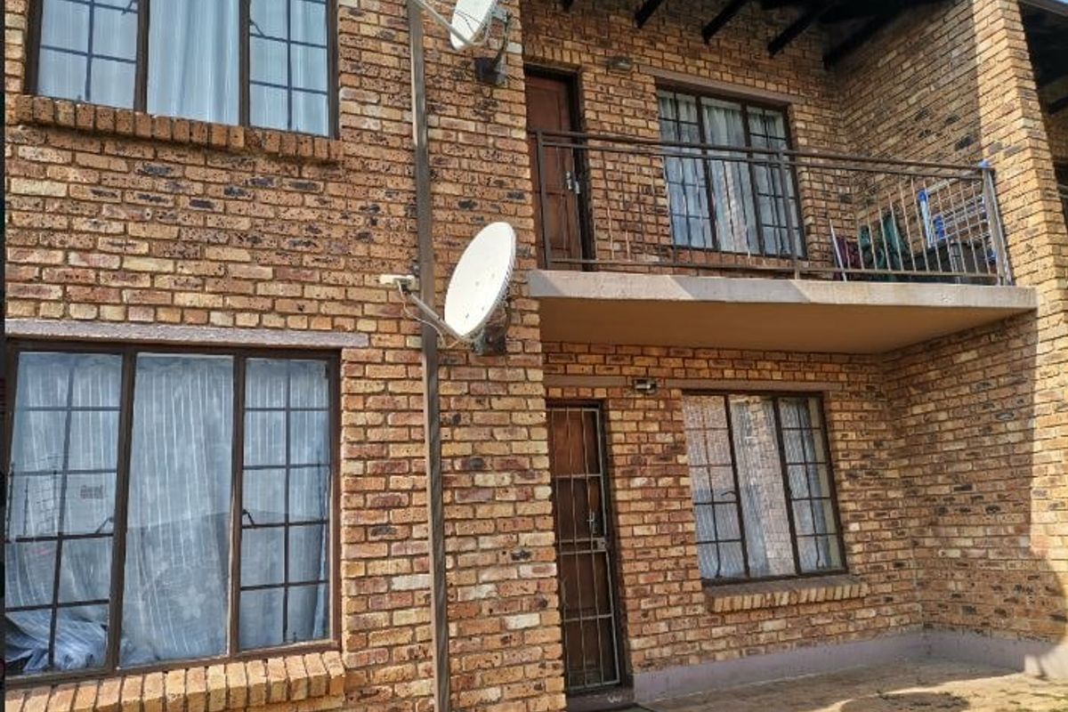 Rear view of a two-story apartment building featuring face brick construction, a private balcony with metal railings, and security gates on the ground floor entrance.