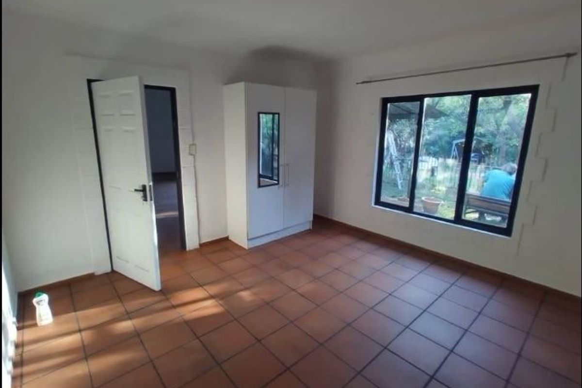 A simple, vacant bedroom featuring terracotta floor tiles, a large window with garden views, and a neutral interior finish.