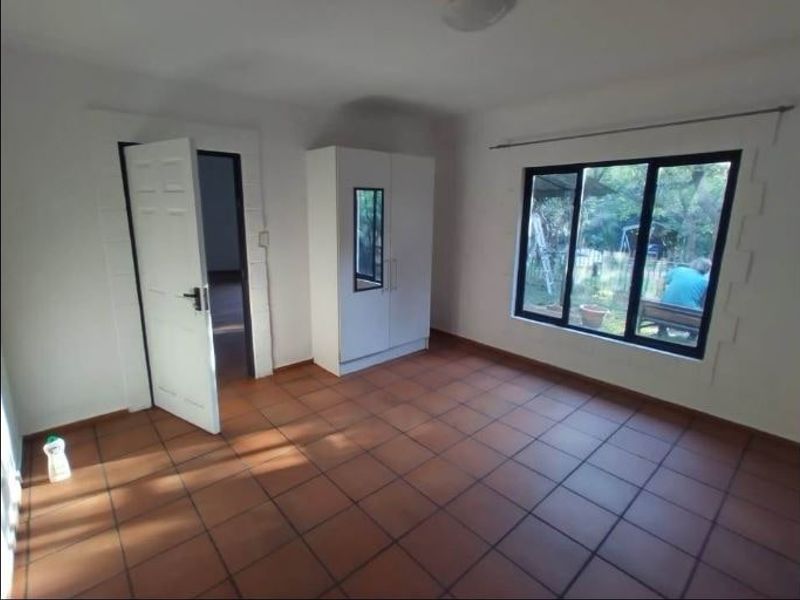 A simple, vacant bedroom featuring terracotta floor tiles, a large window with garden views, and a neutral interior finish.