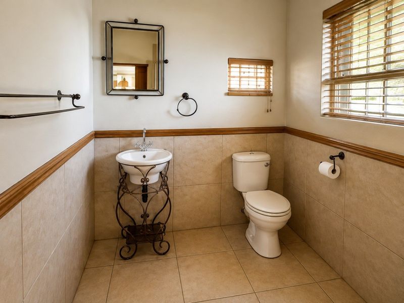 A well-maintained bathroom featuring neutral wall and floor tiling, a pedestal sink, a toilet, and a glass-enclosed shower with wooden window blinds.