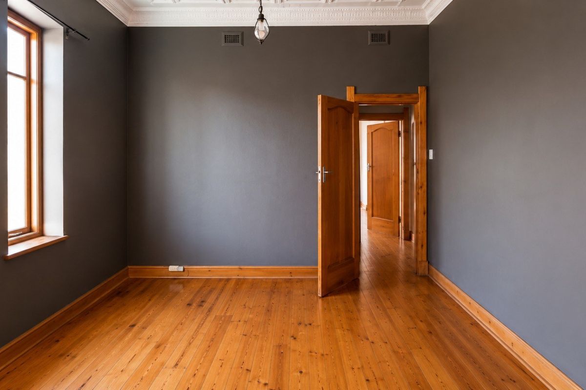 An empty bedroom featuring polished timber flooring, ornate ceiling cornices, and dark grey walls, providing a clean and neutral canvas.