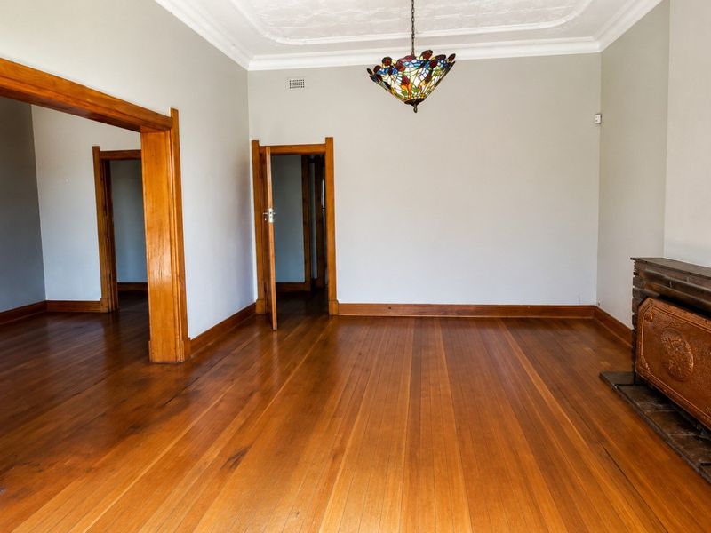 Spacious living room featuring polished hardwood floors, a classic brick fireplace, and original timber door frames with decorative ceiling detailing.