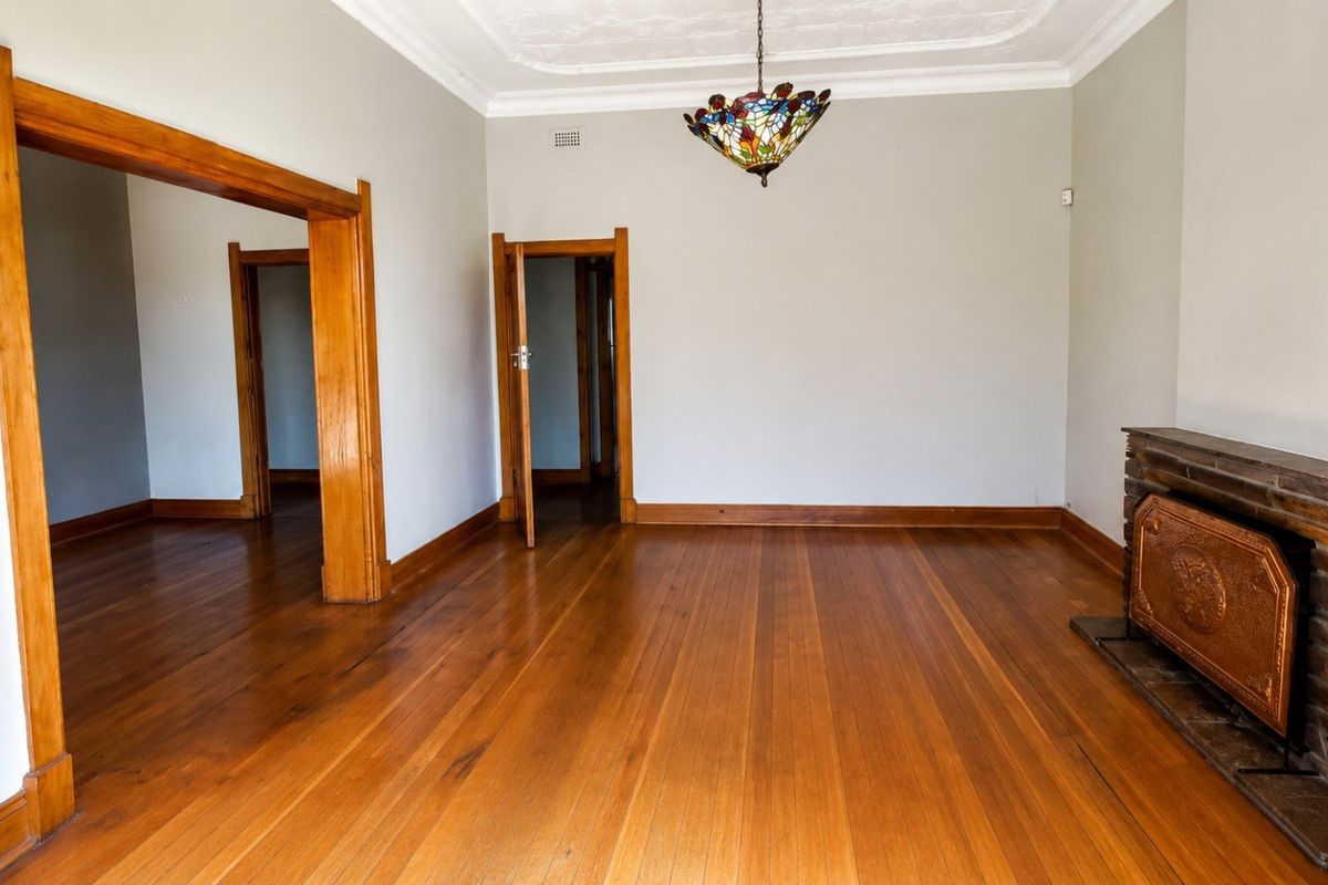 Spacious living room featuring polished hardwood floors, a classic brick fireplace, and original timber door frames with decorative ceiling detailing.