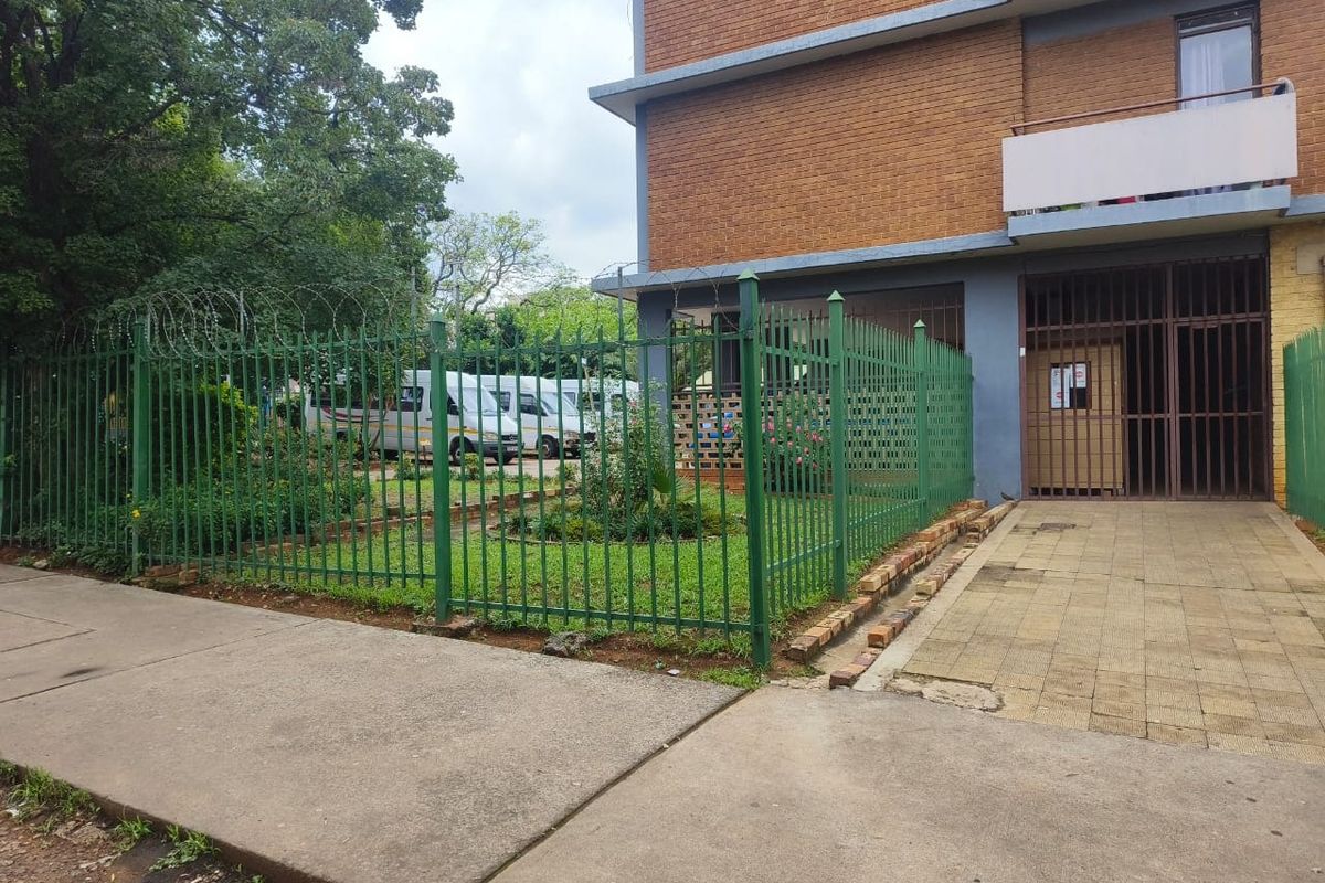 Exterior view of an apartment building featuring a brick facade, balcony, and secure perimeter fencing with a gated entrance.