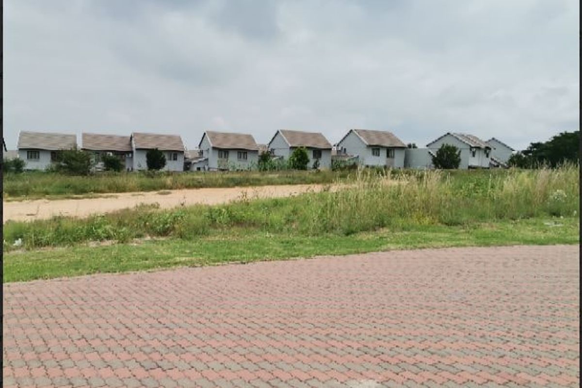 A view of a vacant residential plot featuring a paved driveway and grass verge, with neighboring houses visible in the background.