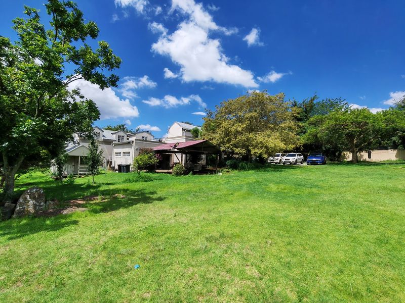 A spacious rear garden featuring a large lawn, mature trees, and a covered patio area attached to the main residential structure.