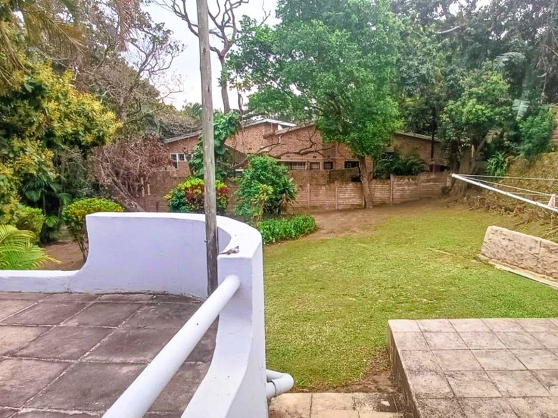 A rear view of the property featuring a tiled patio area, a spacious lawn, and established garden landscaping with a boundary wall.