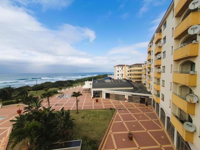 An elevated aerial view of an apartment complex courtyard featuring paved walkways, landscaped gardens, and balconies overlooking the ocean.