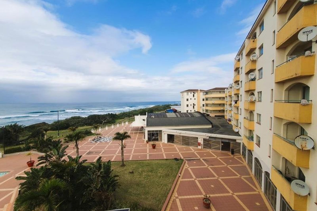 An elevated aerial view of an apartment complex courtyard featuring paved walkways, landscaped gardens, and balconies overlooking the ocean.