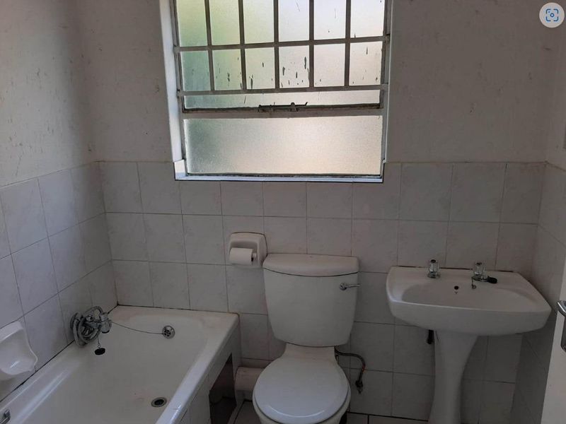 A dated bathroom featuring a standard bathtub, pedestal basin, and toilet, with tiled walls and a security-barred window.