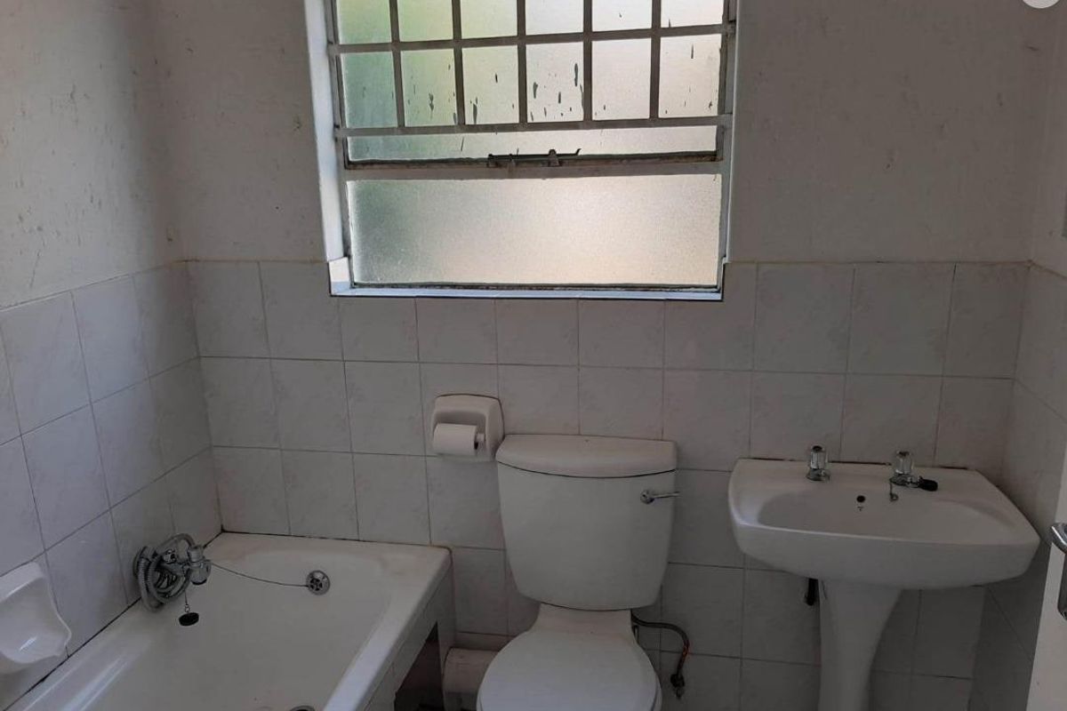 A dated bathroom featuring a standard bathtub, pedestal basin, and toilet, with tiled walls and a security-barred window.