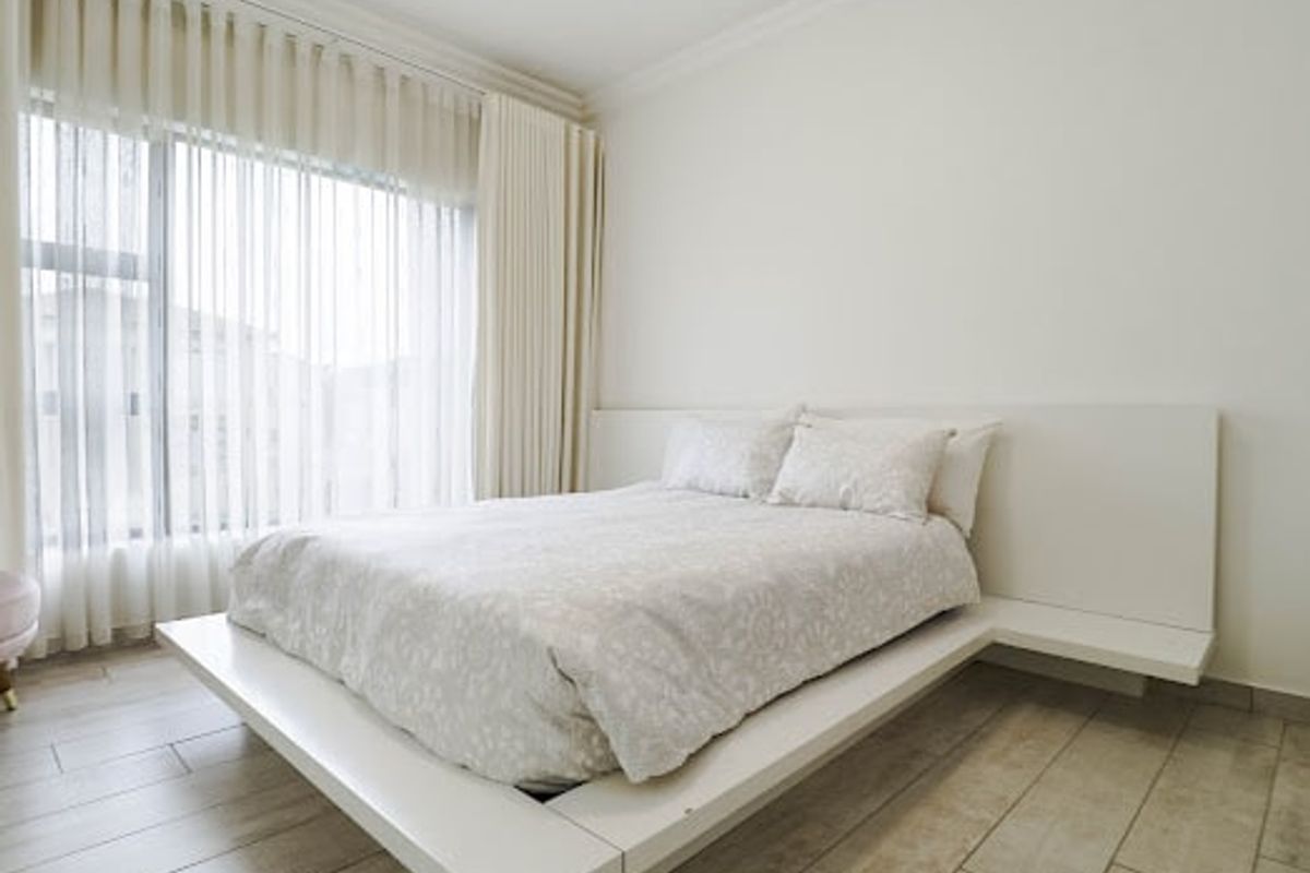 A bright and modern bedroom featuring a built-in platform bed frame, neutral tiled flooring, and floor-to-ceiling window treatments.