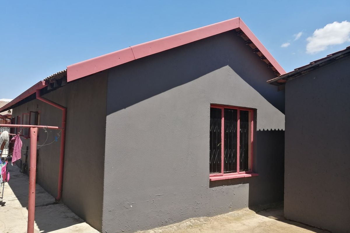 Rear exterior view of the house featuring grey rendered walls, red-painted roof trim and window frames, and a concrete pathway.