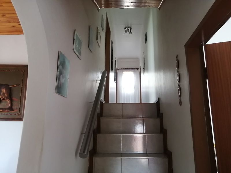 A tiled staircase hallway featuring a metal handrail and timber ceiling accents, leading to an upper level with natural light.