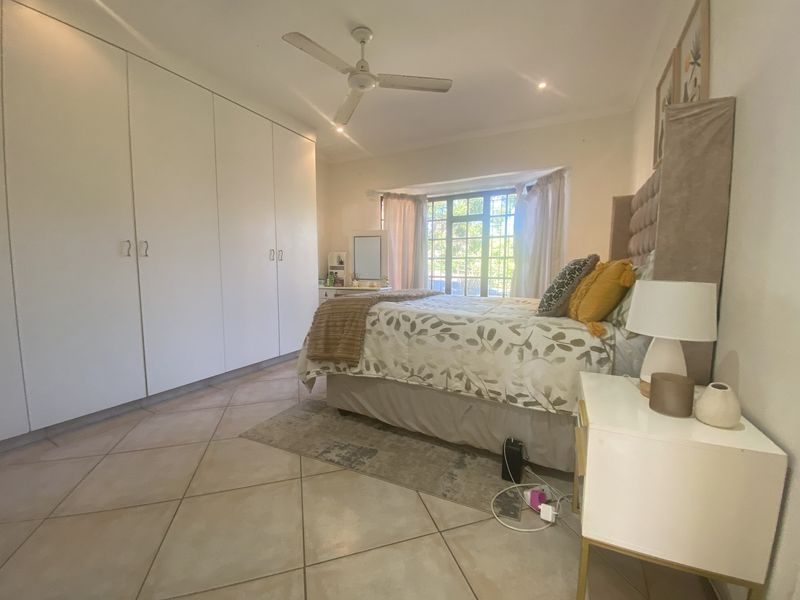 A well-lit bedroom featuring extensive built-in cupboards, tiled flooring, a ceiling fan, and a large bay window allowing for natural light.