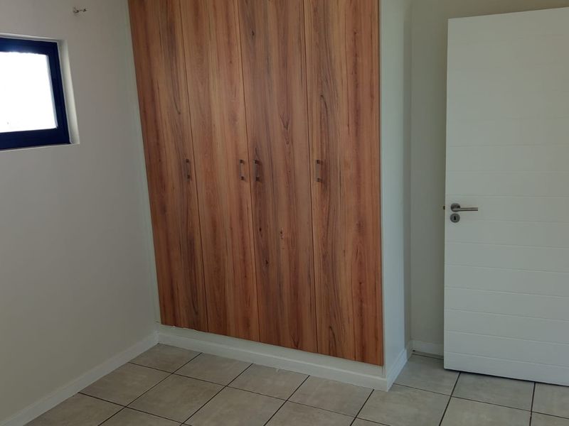 A clean, empty bedroom featuring modern wood-finish built-in cupboards, neutral tiled flooring, and a sliding window.