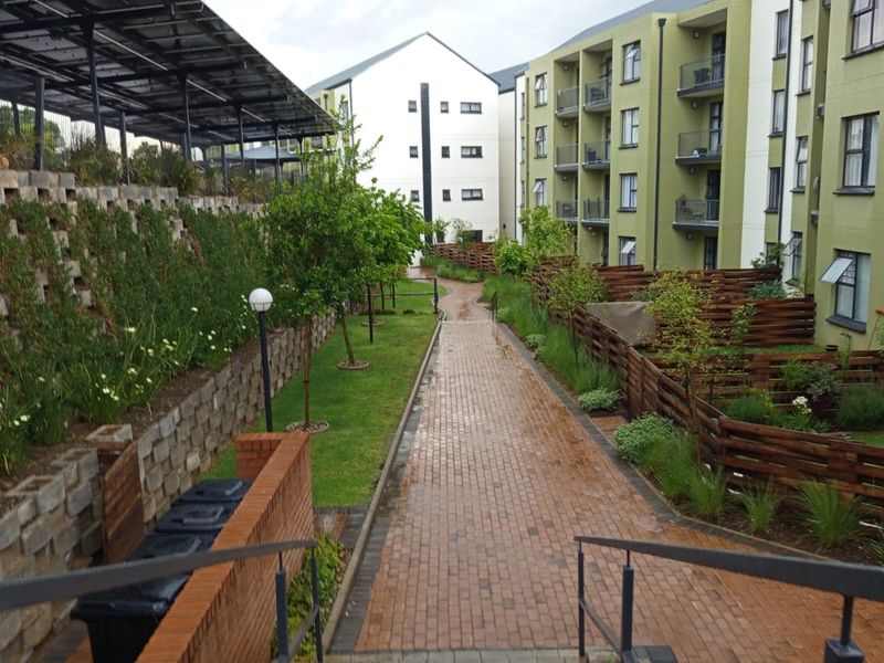 A well-maintained communal garden area featuring a paved walkway, timber fencing, and retaining walls between apartment blocks.