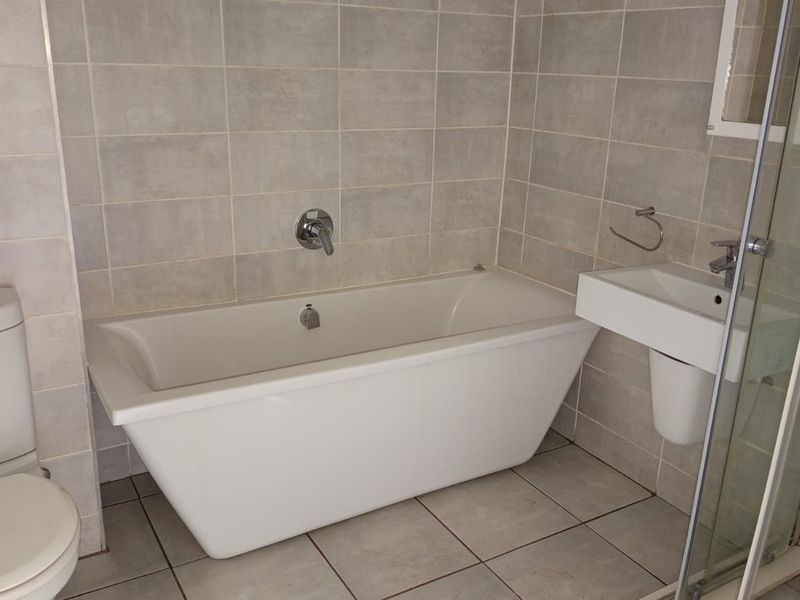 A clean, modern bathroom featuring a white bathtub, wall-mounted basin, toilet, and a glass-enclosed shower with neutral ceramic tiling throughout.