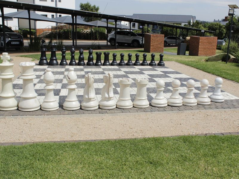 Outdoor communal garden area featuring a large paved chess board, manicured lawn, and adjacent covered parking facilities within the apartment complex.