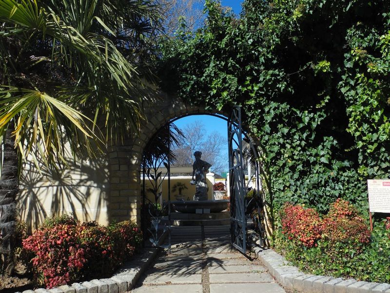 A charming garden entrance featuring a stone archway, wrought iron gate, and a central stone fountain surrounded by established landscaping.