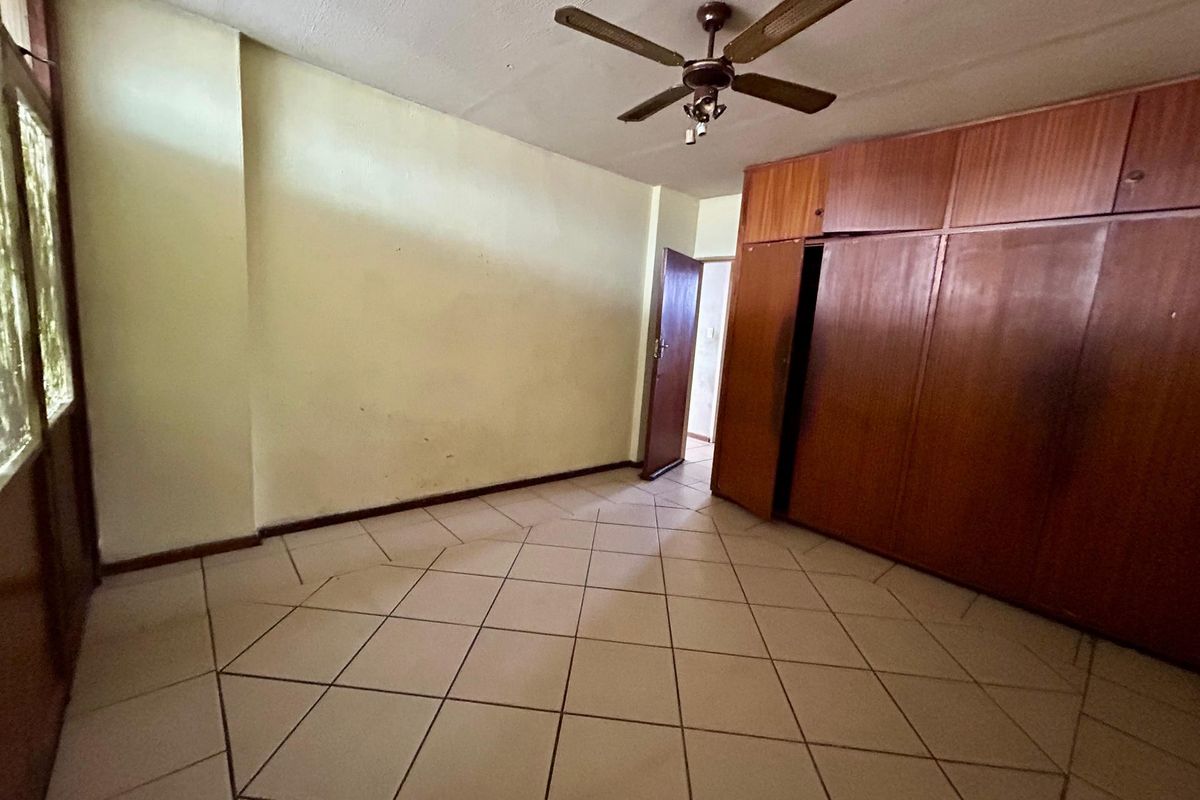 An empty bedroom featuring tiled flooring, a ceiling fan, and extensive built-in wooden cupboards, offering a blank canvas for renovation.