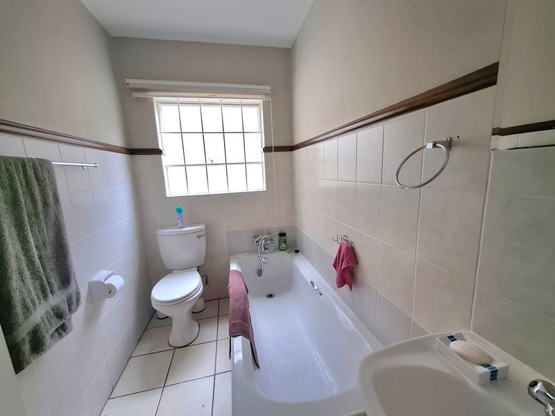 A functional bathroom featuring a bathtub, toilet, and washbasin with tiled finishes and a window with security bars.