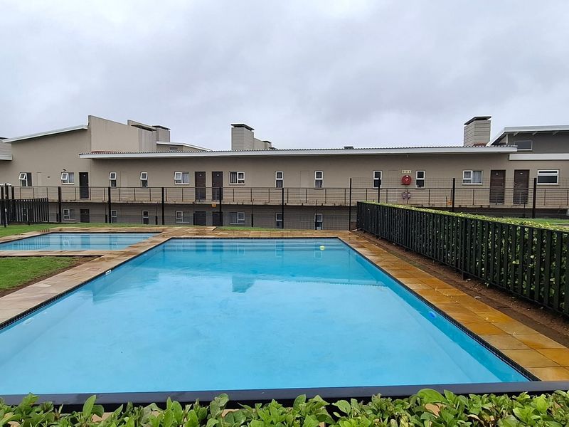 Communal swimming pool and wading pool area featuring paved surrounds, manicured lawn, and secure metal perimeter fencing within the apartment complex.