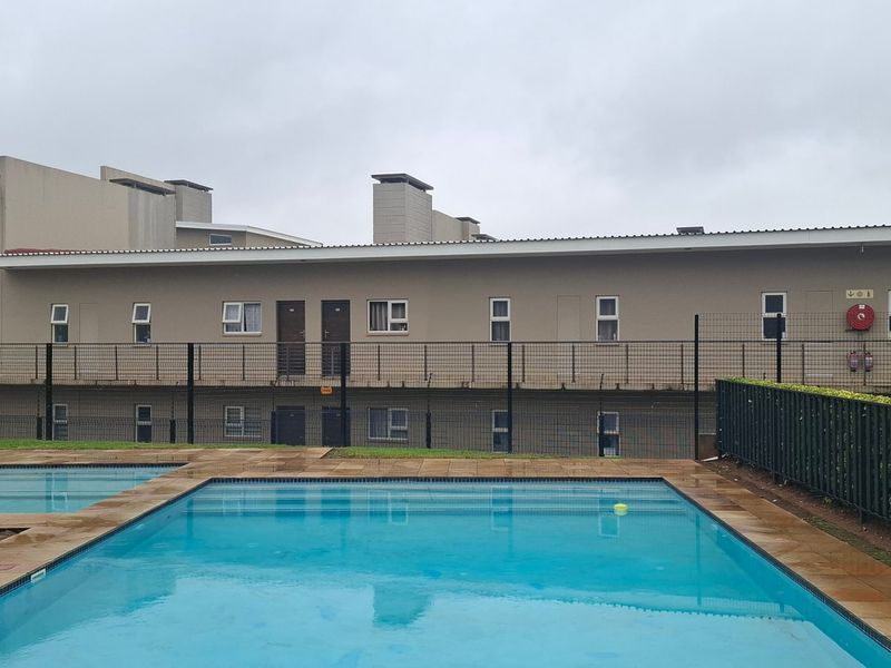 A communal swimming pool area featuring a paved deck and perimeter fencing, set against the exterior facade of the apartment complex.