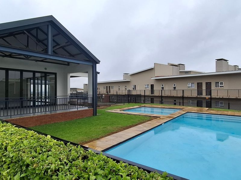 A well-maintained communal pool area featuring a paved deck, landscaped lawn, and a covered patio structure with modern architectural finishes.