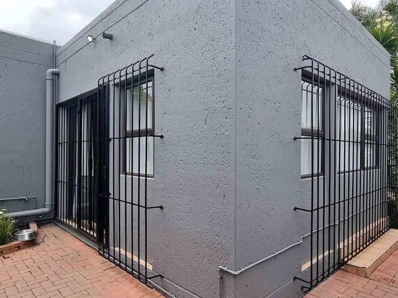 Rear exterior view of the cluster property featuring a textured grey facade, security bars on windows and doors, and a paved patio area.