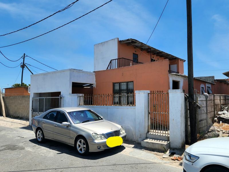 Front elevation of a two-story house featuring a balcony, metal fencing, and a concrete boundary wall, showing signs of significant exterior wear.