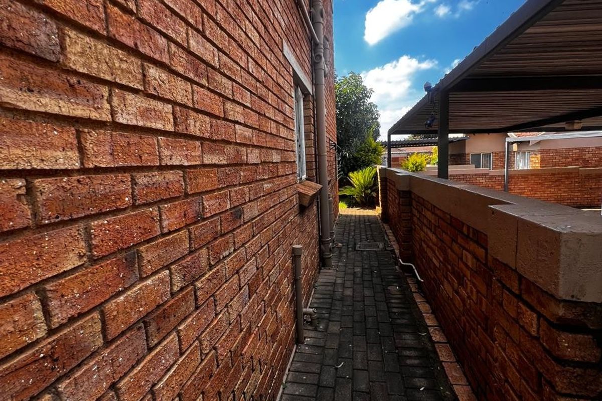 A narrow paved side walkway between a brick building facade and a boundary wall, featuring a covered carport structure.